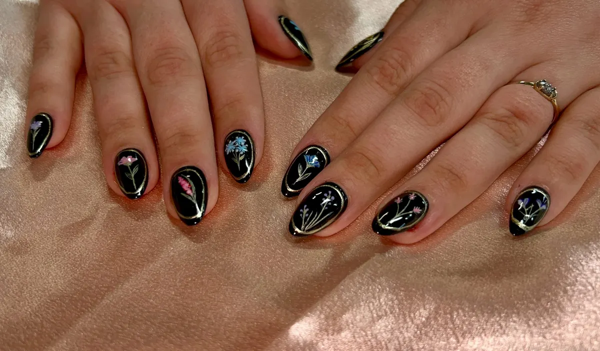 Glossy black gel nails with gold-framed pressed wildflowers, by Victoria
